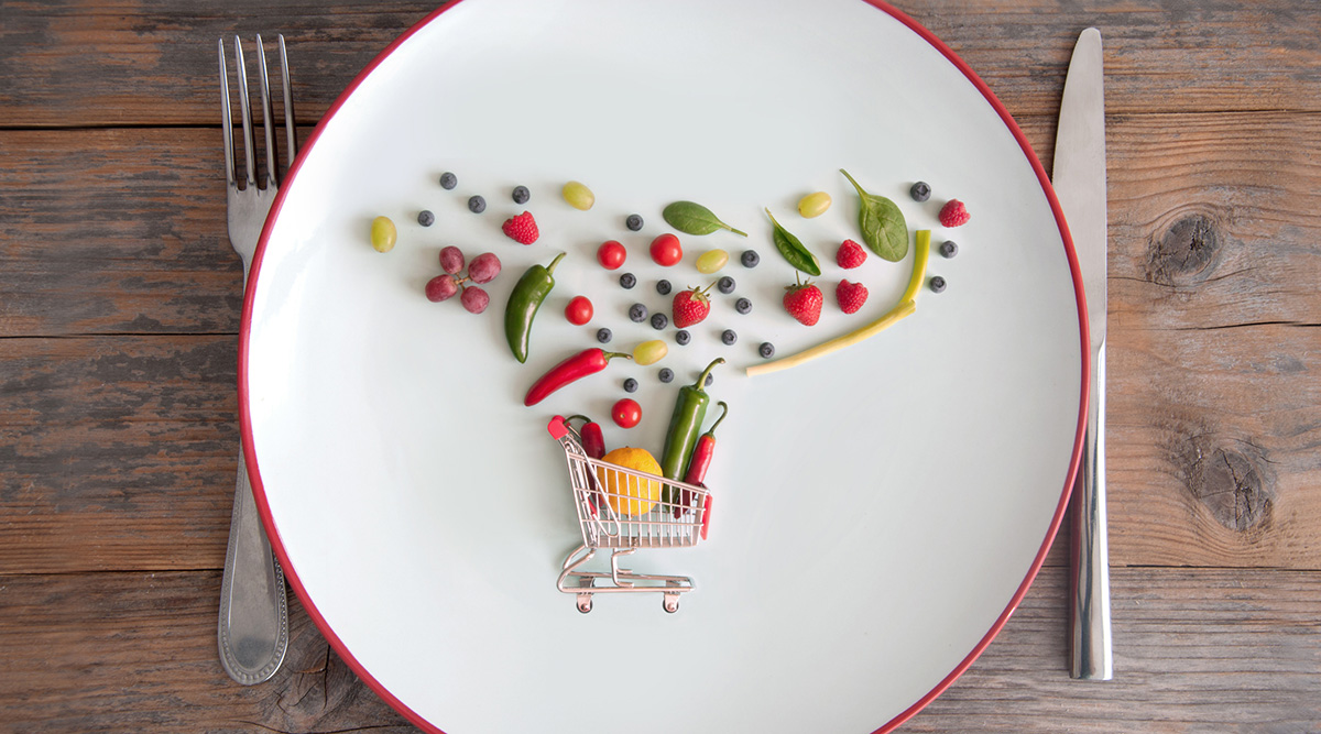 A tiny grocery cart and tiny produce dispersed on a plate with a fork and knife on either side.