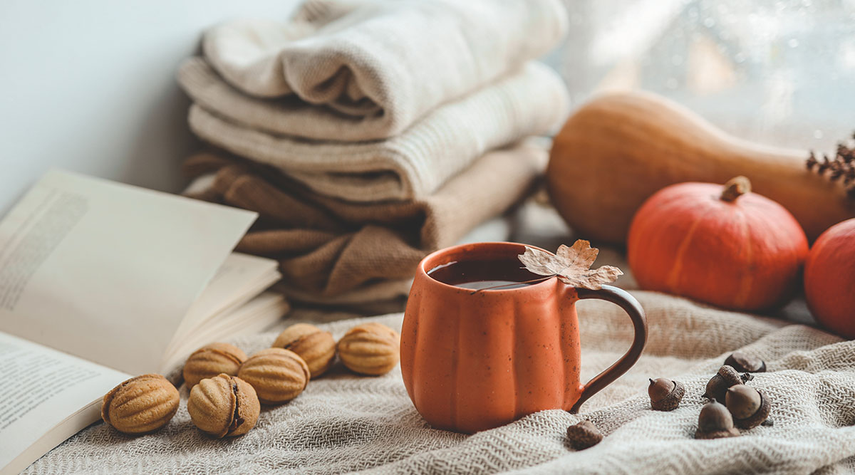 A pumpkin mug filled with hot cocoa surrounded by walnuts, acorns, pumpkins, a squash, and a book.