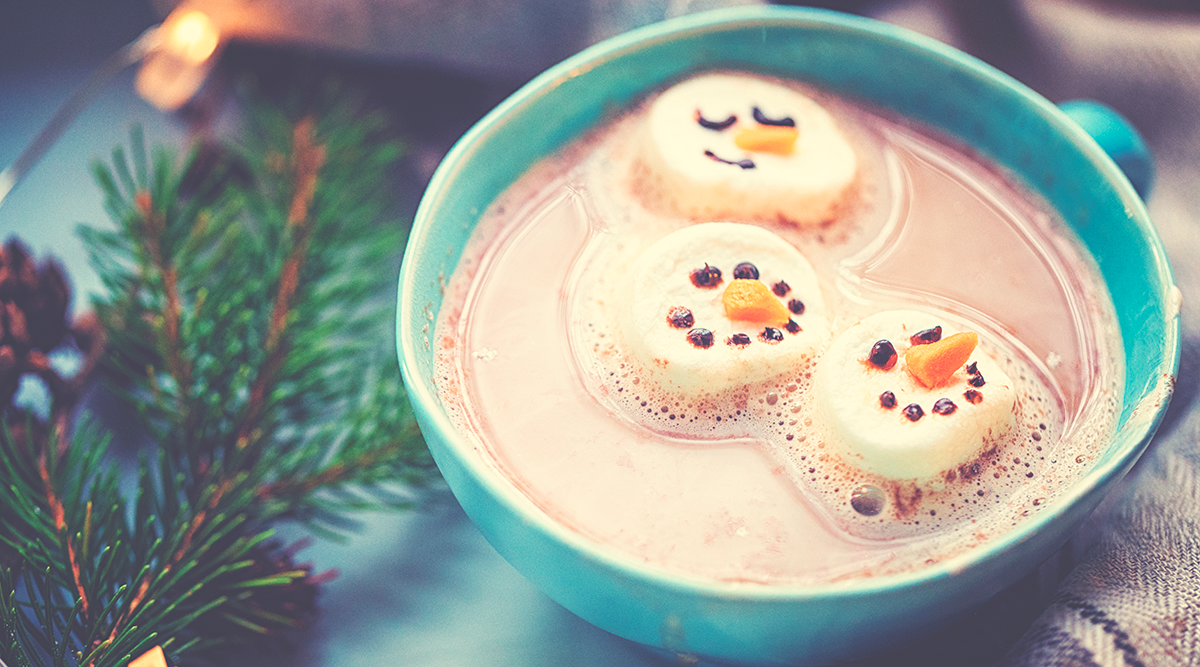 Hot cocoa with snowmen faces on marshmallows in a blue mug next to a pine branch